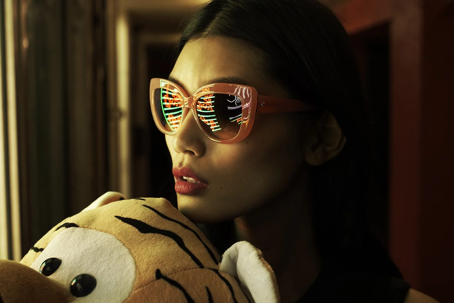 A woman wearing pink sunglasses with reflections of neon lights, holding a plush tiger toy, inside a dimly lit room. Advertising. Producer. Miranda Kendrick. Kate Spade.