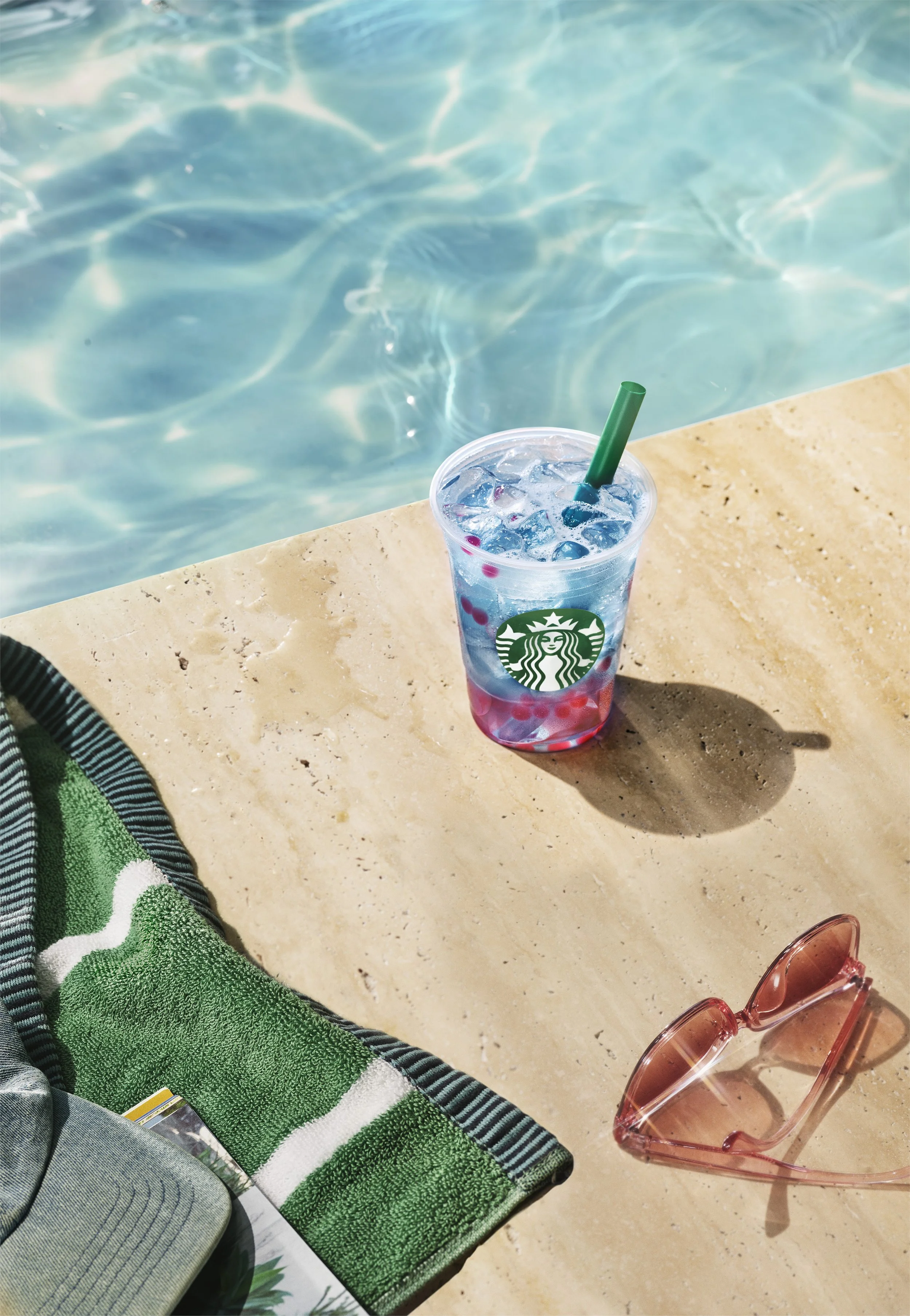 A close-up of a Starbucks cup filled with a clear beverage and ice, resting on a beige surface near a swimming pool with bright sunlight reflecting on the water.