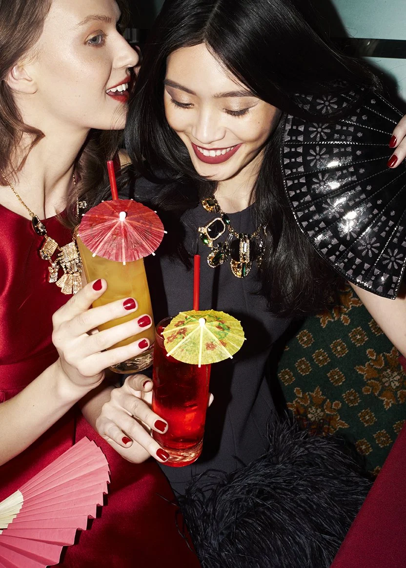 Two women celebrating with colorful cocktails topped with paper umbrellas, holding drinks together, dressed festively, with one holding a decorative black fan.