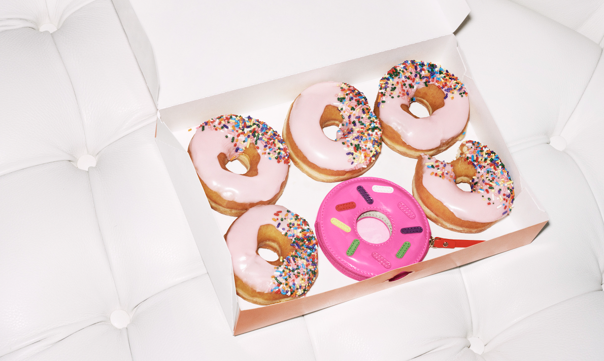 A white box containing five donuts with pink icing and colorful sprinkles, and a pink plastic toy donut with sprinkles, all on a white tufted surface.