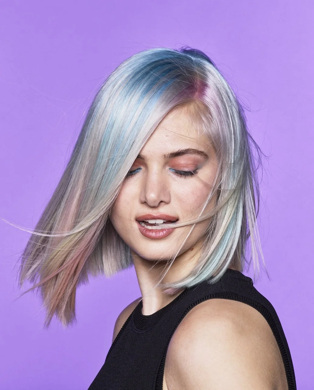 A woman with pastel-colored hair in shades of silver, pink, blue, and purple, styled in a shoulder-length bob, against a lavender background. She is wearing a black sleeveless top and has her eyes closed, with a slight smile and a hand touching her n