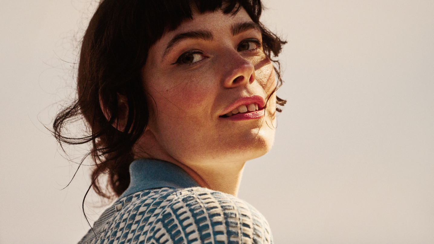 A woman with short dark hair smiling, looking over her shoulder, wearing a light blue and white knitted sweater, with a neutral background.