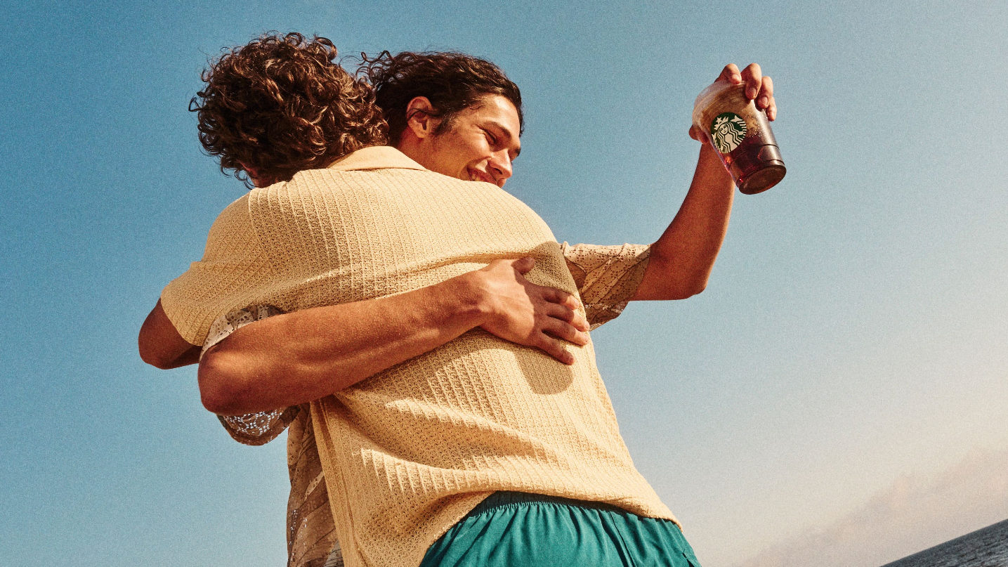Two young men hugging at the beach under a clear blue sky, one holding a Starbucks iced coffee, smiling