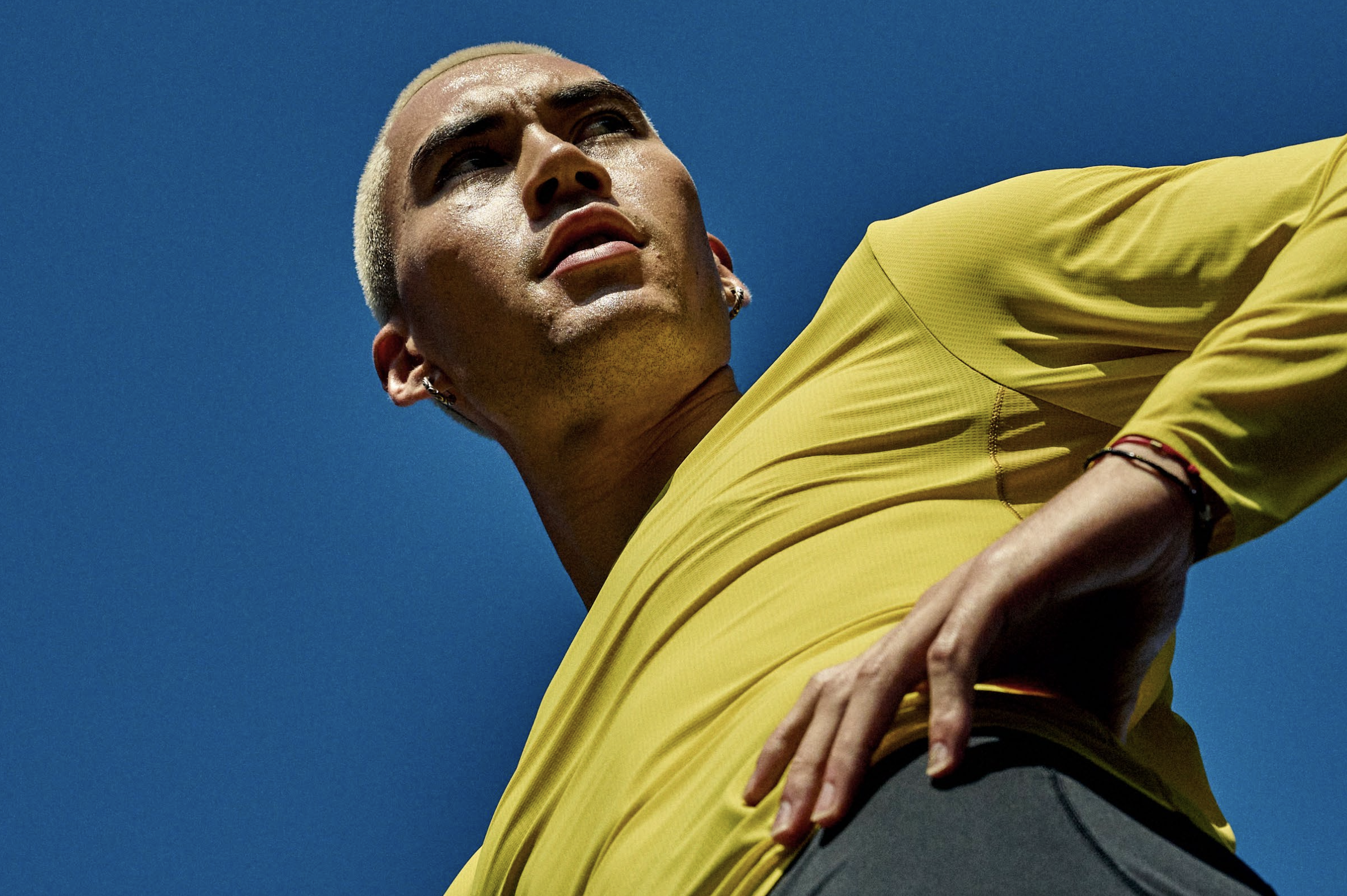 Close-up of a person with short platinum blonde hair, wearing a yellow athletic shirt, posing against a clear blue sky. Advertising. Producer. Miranda Kendrick. HOKA.