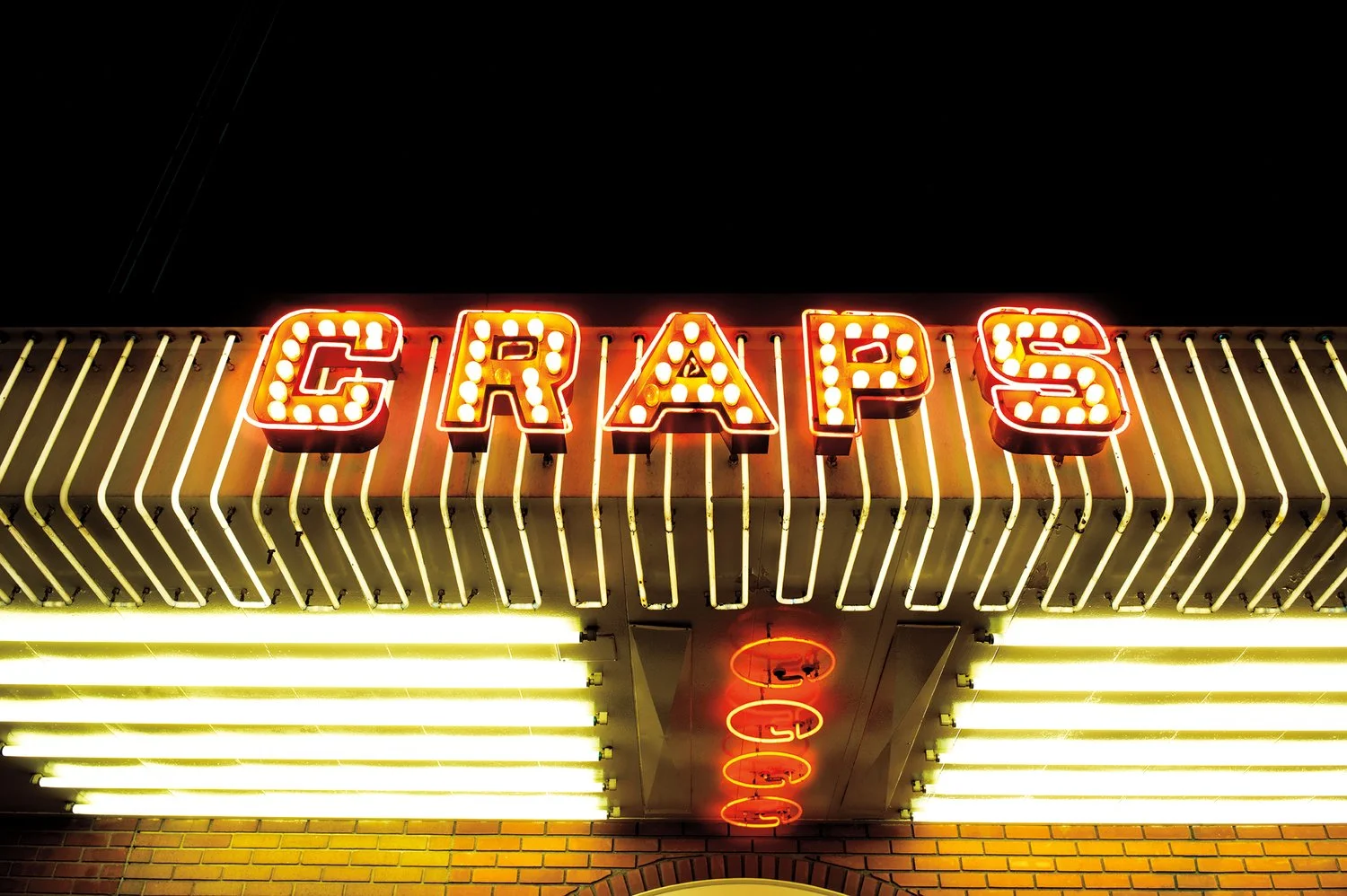 Neon sign spelling out 'CRAPS' with bright bulbs, above a row of yellow fluorescent lights and a brick wall. Vintage Las Vegas.
