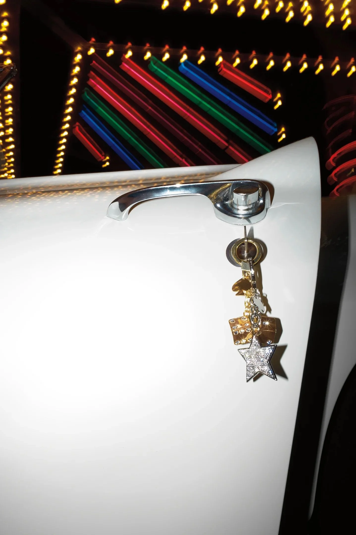 Close-up of a white surface with a metallic handle and a charm hanging from it, featuring a star and chip design, with colorful neon or LED lights in the background forming diagonal lines. Kate Spade. Las Vegas.
