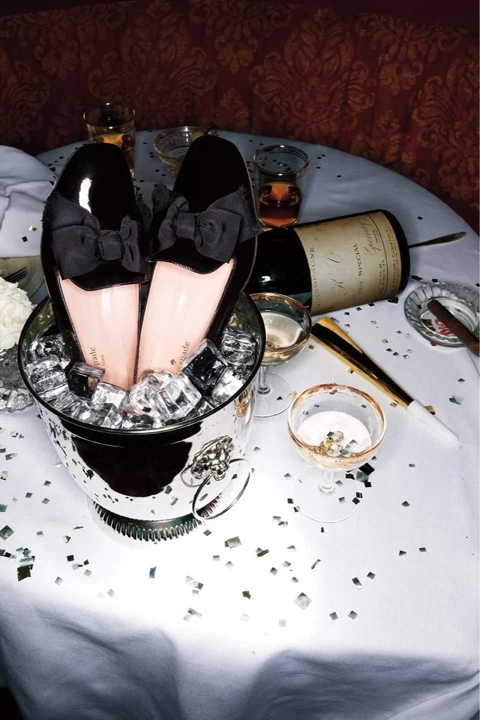 A table set with champagne, glasses, and an ice bucket containing a bottle and a pair of shoes with bows, decorated with confetti.