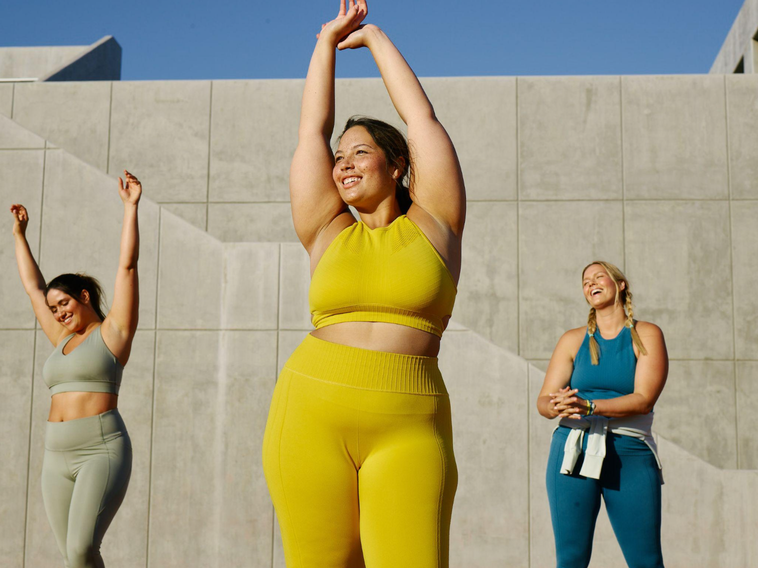 Three women doing yoga outdoors in front of a concrete wall, smiling and stretching with arms raised, wearing colorful athletic clothing on a sunny day.