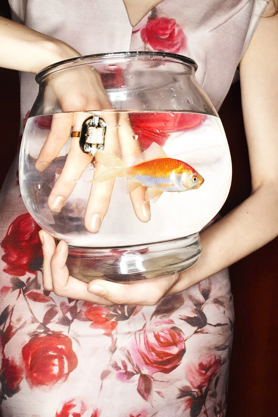 Person holding a clear fishbowl with a goldfish inside, wearing a ring and a floral dress.