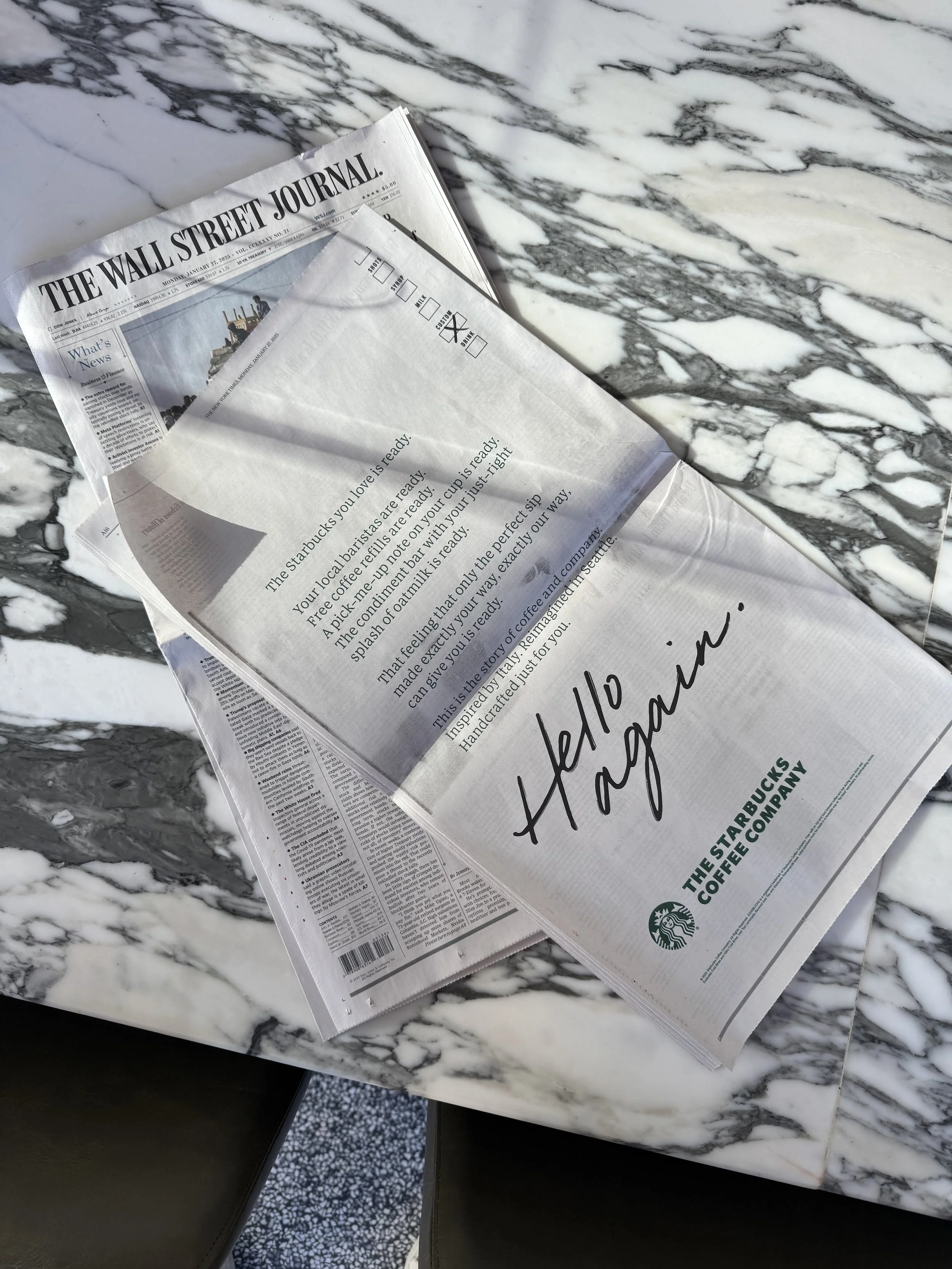 A newspaper on a marble table with the headline 'The Wall Street Journal' and a Starbucks advertisement that says 'Hello again'.