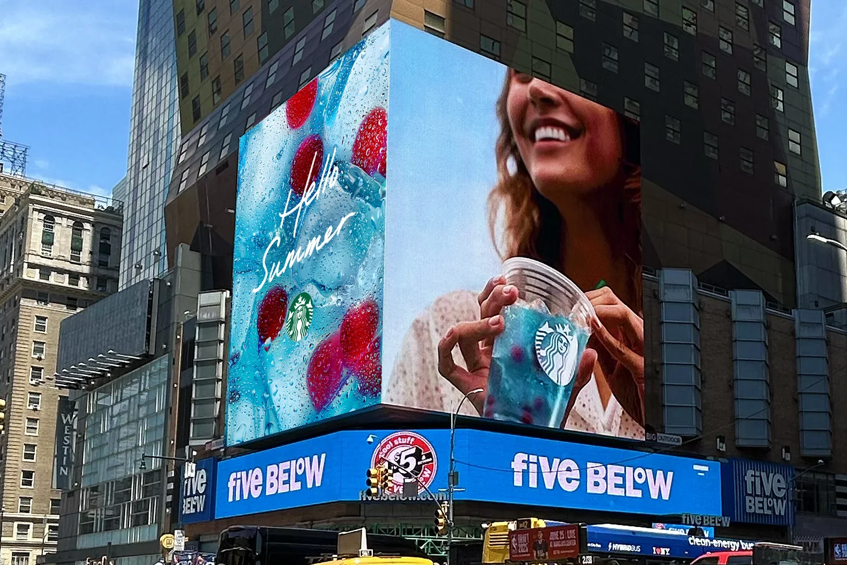 An electronic billboard in Times Square, New York City, featuring an ad for Starbucks Hello Summer beverages, with a woman smiling and holding a colorful drink with the Starbucks logo, surrounded by tall buildings and city infrastructure.