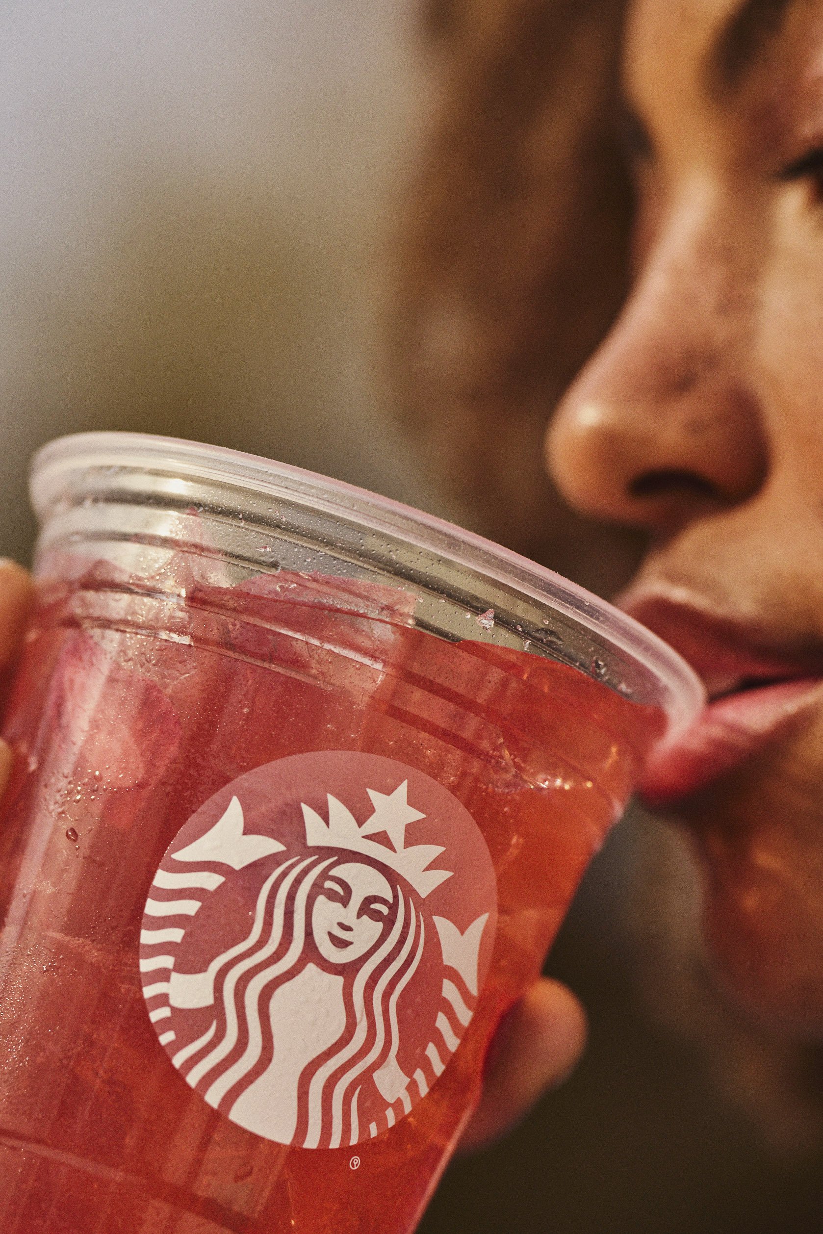 Close-up of a person's face in profile drinking a pink Starbucks beverage from a clear plastic cup with a white logo, possibly a smoothie or frappuccino. Advertising. Producer. Miranda Kendrick.