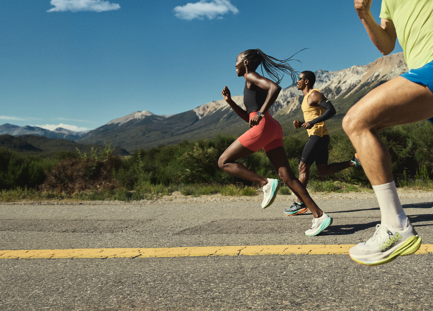 Group of runners running on a paved road outdoors with mountains in the background under clear blue sky.