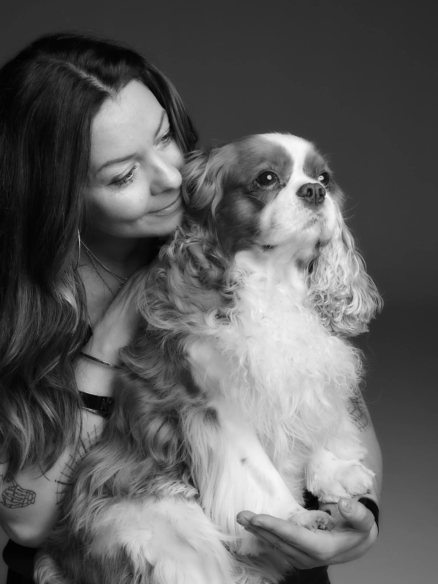 A woman with long dark hair smiling and holding a Cavalier King Charles Spaniel dog against a plain background.