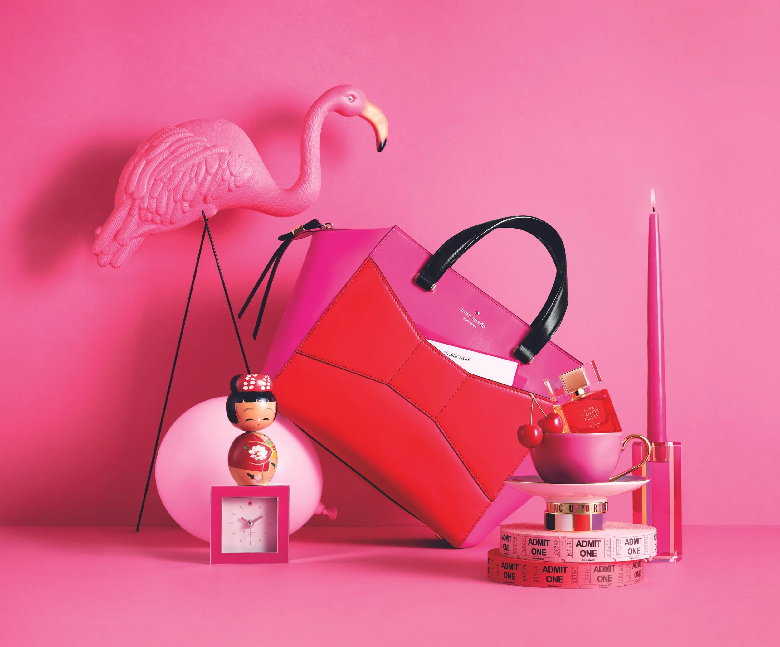 Pink background with a pink flamingo figurine, pink purse, pink clock, Japanese dolls, pink candle, decorative rings, and tickets stacked on a table.