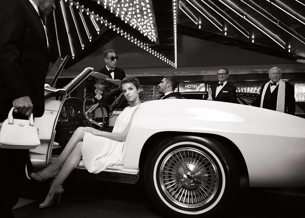 Black and white photo of a woman in a white dress sitting in a vintage convertible car surrounded by five men dressed in tuxedos in a neon-lit setting. Anna Kendrick. Las Vegas. Kate Spade.