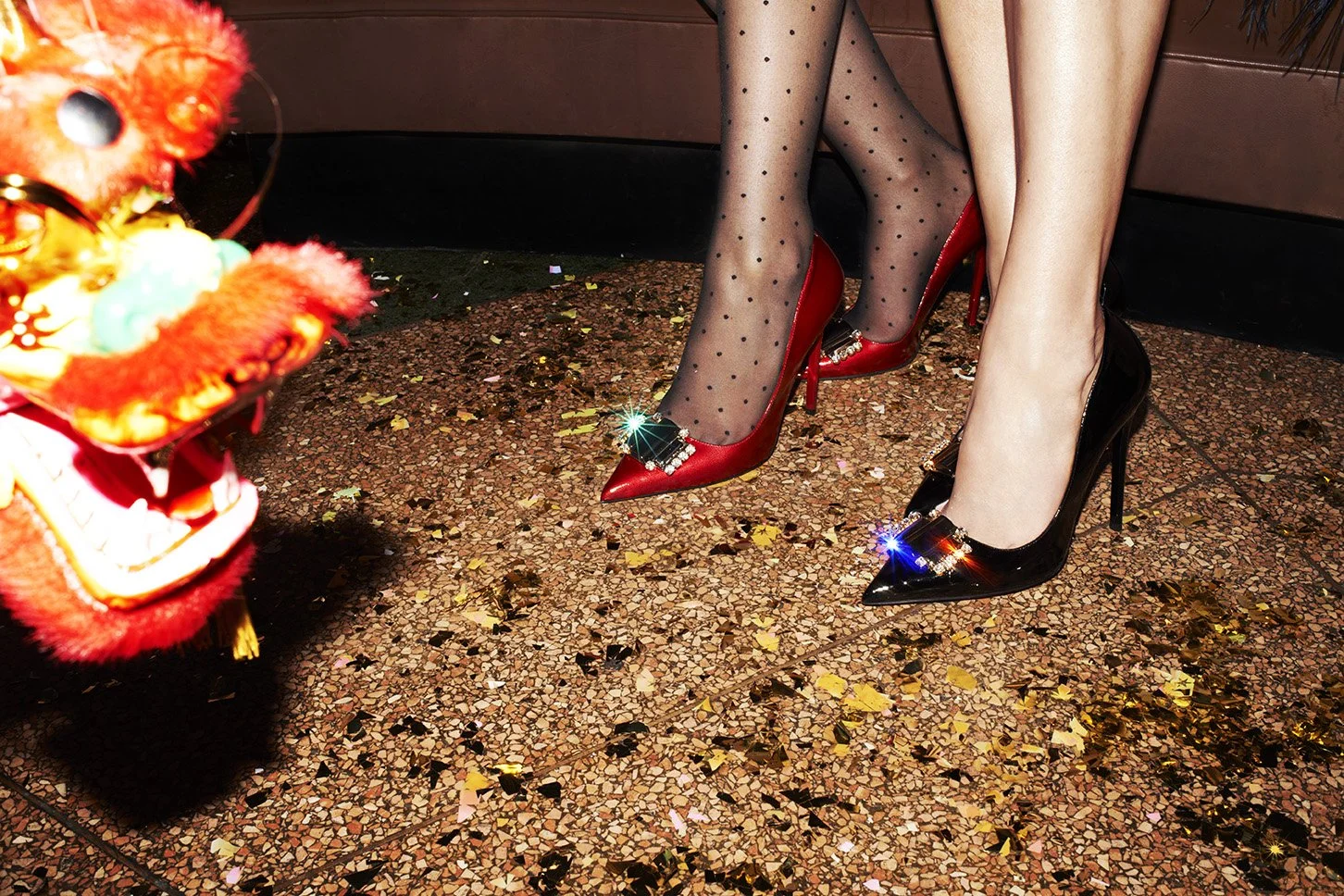 Two women wearing high-heeled shoes, one in red and one in black, standing on a glittery floor with scattered confetti. One woman has polka dot stockings, and the other has bare legs. A colorful lion dance costume is visible on the left.