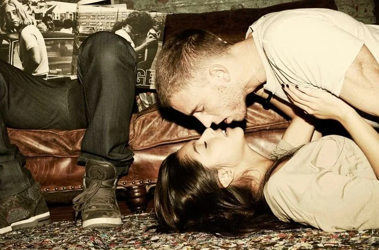 A man and woman are lying on the floor facing each other. The man is leaning over the woman, and they are about to kiss. . There is a sofa and another person reads Joseph Szabo Teenage photography book. Advertising. Producer. Miranda Kendrick. Axe.