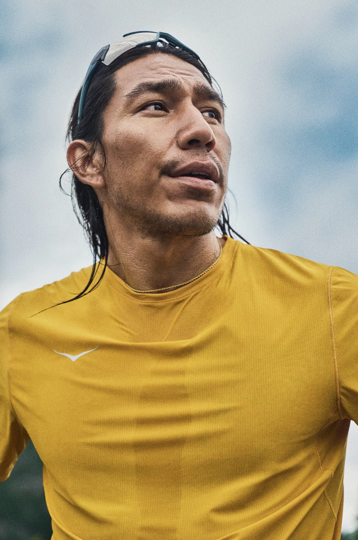 A close-up of a man wearing a yellow sports shirt, with sunglasses resting on his head, looking into the distance against a cloudy sky. HOKA. Advertising. Producer. Miranda Kendrick. Anthony Blasko.