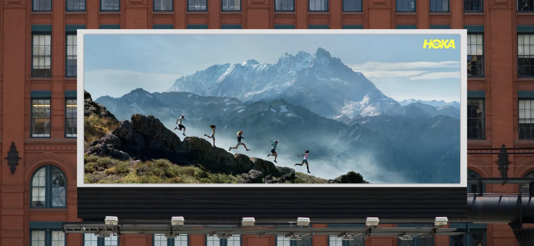 A billboard on a brick building showing a mountain landscape with snow-capped peaks and a group of six people running and jumping across rocks in the foreground.