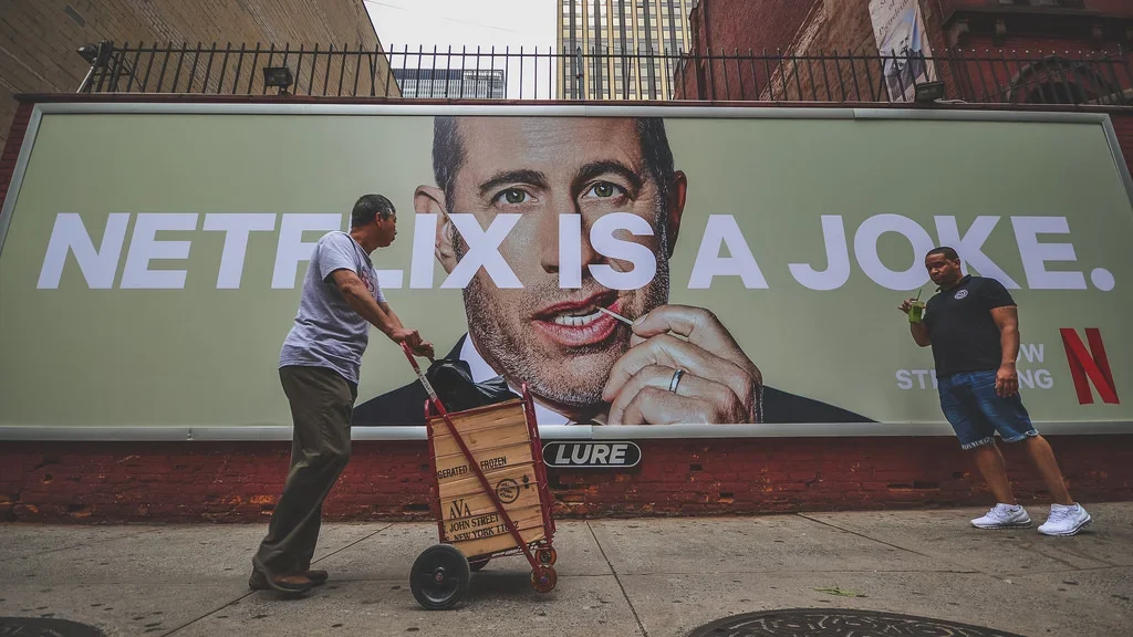 The billboard features a large image of david chapelle and contains the text 'NETFLIX IS A JOKE.'  Advertising. Producer. Miranda Kendrick.