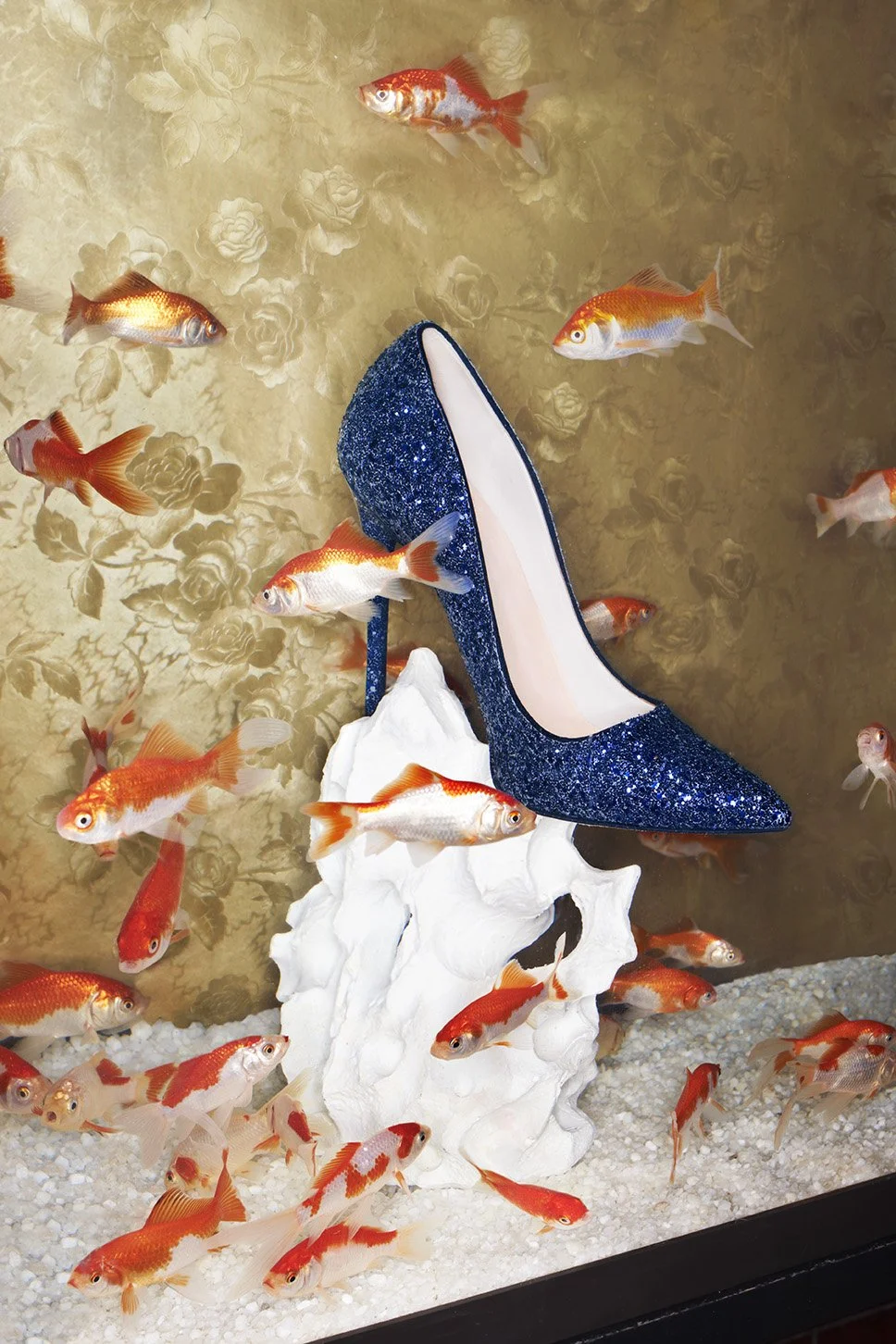 A decorative display in an aquarium with orange and white goldfish swimming around a high-heeled shoe covered in blue glitter, resting on a white artificial rock, against a floral-patterned background.
