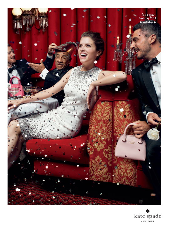 Group of people celebrating at a party with red curtains, confetti, and elegant attire. Anna Kendrick a sparkly dress and men in tuxedos, some holding handbags and one wearing a party hat. Kate Spade.
