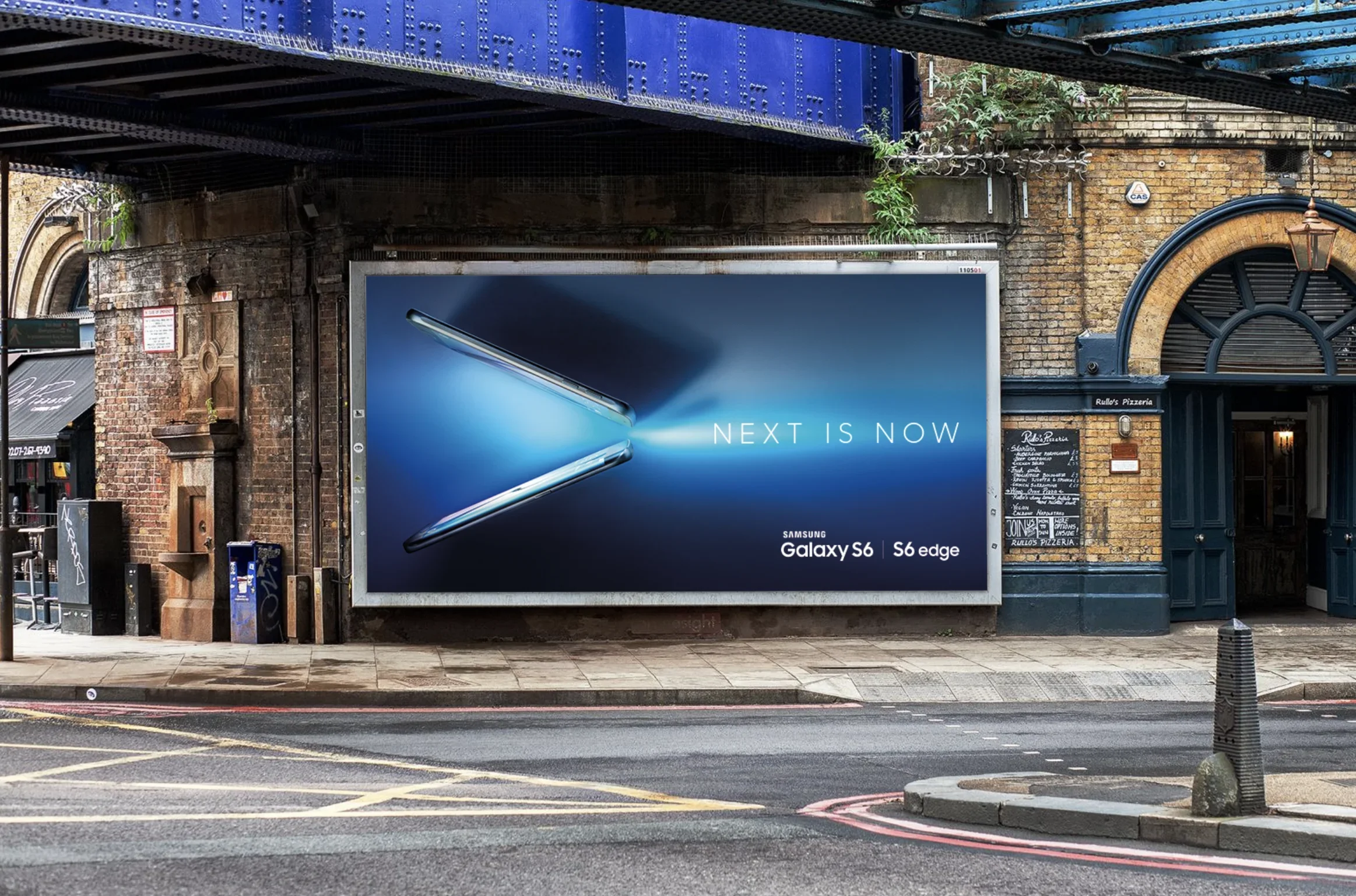 An urban street scene with a large digital billboard advertising the Samsung Galaxy S6 and S6 Edge, featuring a close-up image of the phones and the slogan "NEXT IS NOW." Surrounding the billboard are brick buildings, storefronts, and street furniture, with some greenery and a blue bridge overhead.