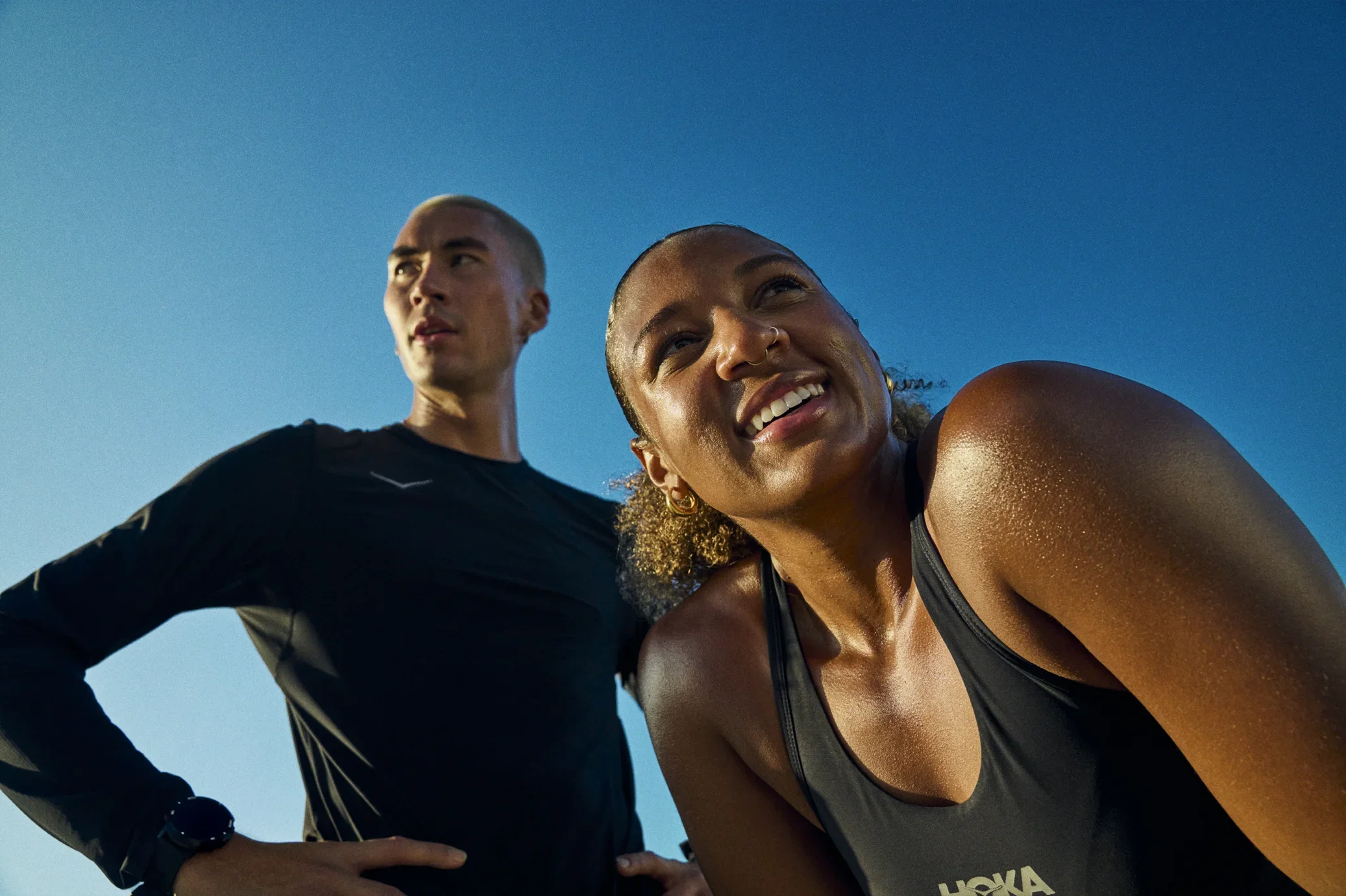 Two diverse women and a man in athletic wear outdoors under a clear blue sky, smiling and looking happy, possibly after exercise. Athletes. Running. HOKA.