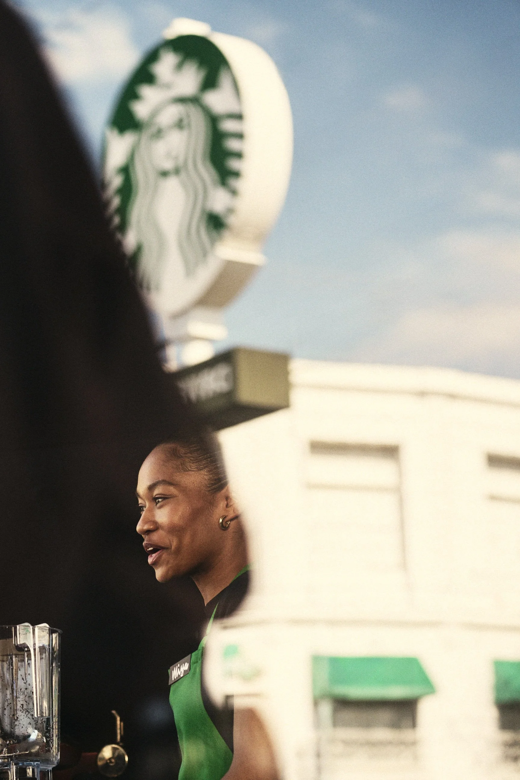 Barista smiling, wearing a black and green starbucks uniform, seen through drive through window. Advertising. Producer. Miranda Kendrick. Starbucks.