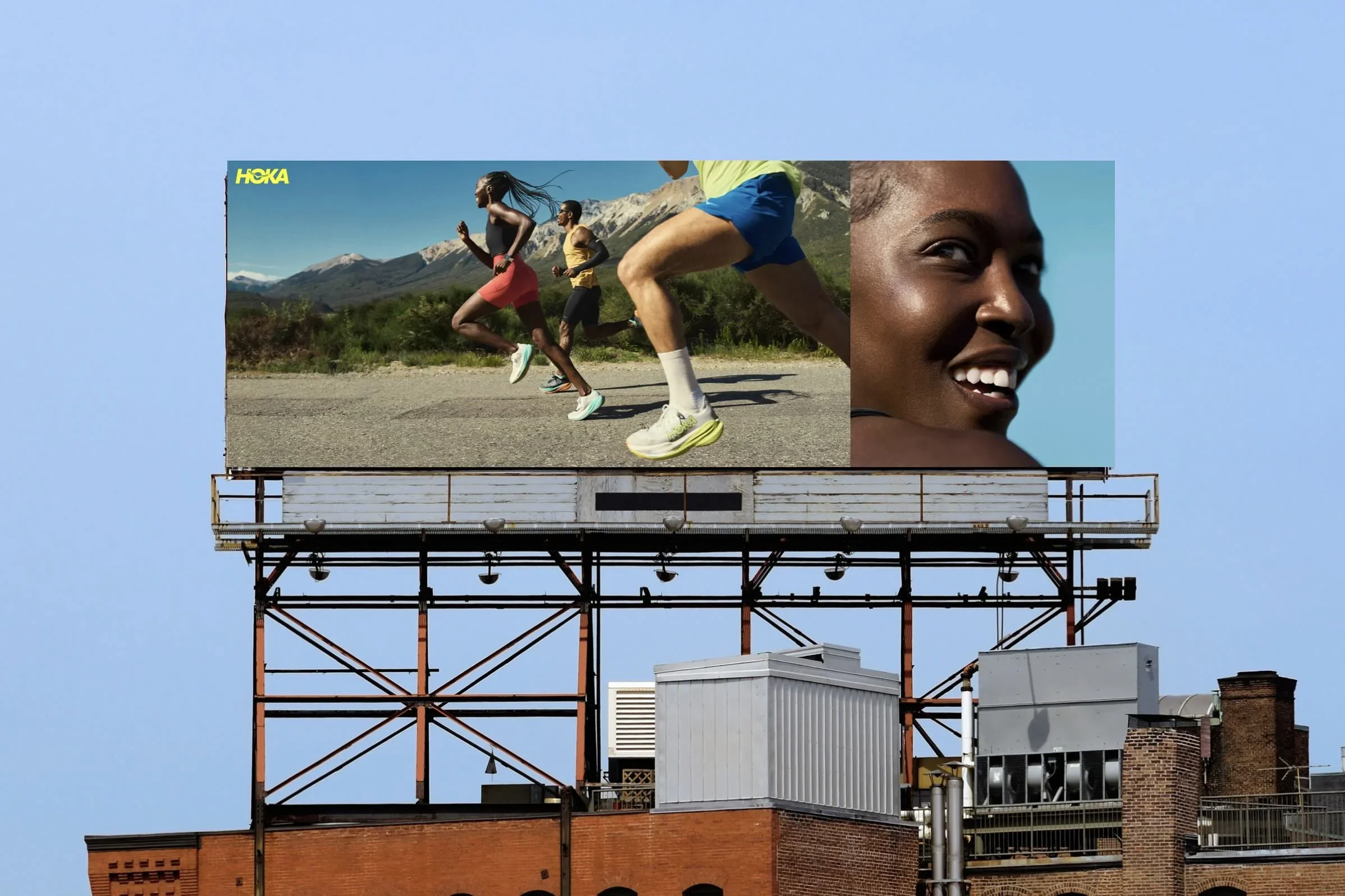 Billboard on a building featuring a photo of three runners, two men and one woman, jogging outdoors with mountains in the background; a smiling woman close-up on the right side of the billboard. HOKA. Runners. Advertising. Producer. Miranda Kendrick.