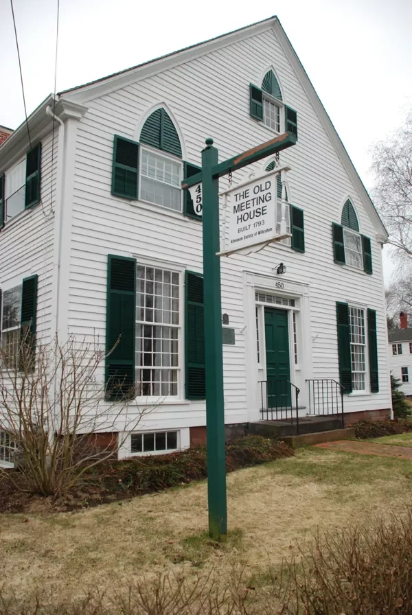 A white historic building with green shutters and a sign that reads "The Old Meeting House, Built 1793," located on a residential street with a lawn in front.