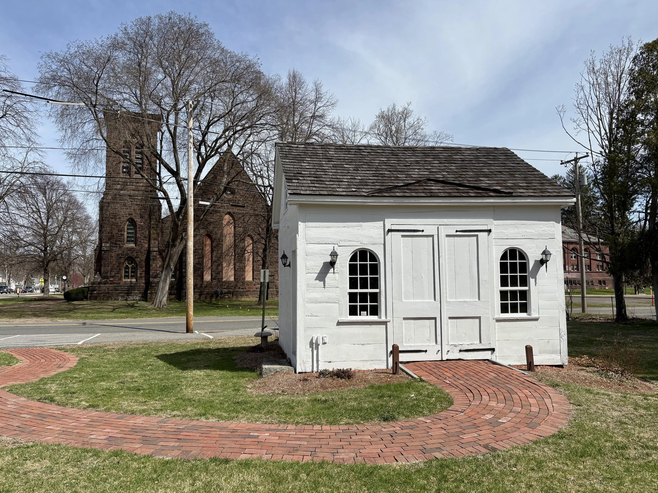 A small white wooden building with black lantern-style lights on either side of the door and arched windows. In front is a curved brick pathway on a lawn, with a large historical church or old building with stone walls and tall windows in the background, along with leafless trees.