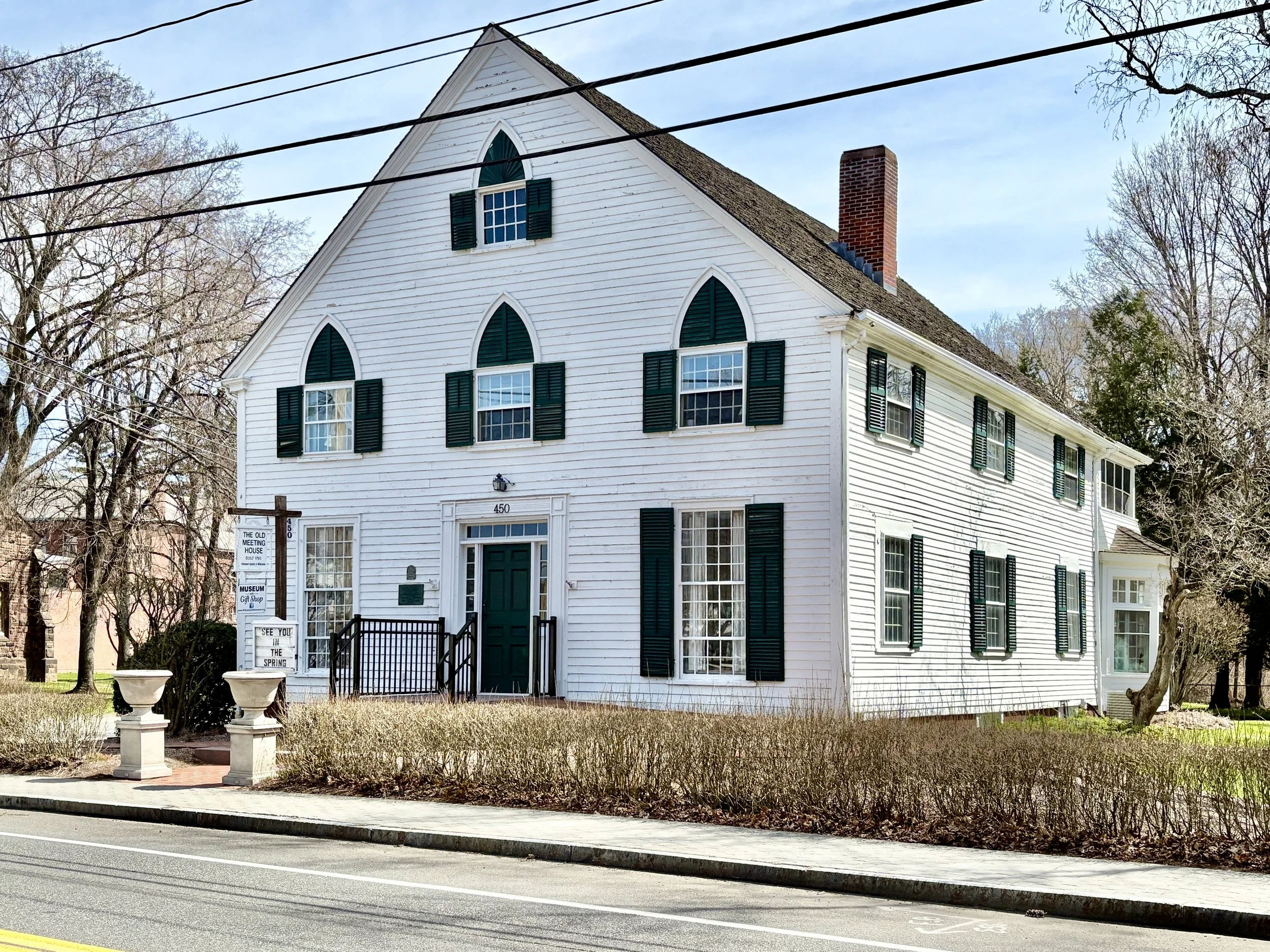 A white, two-story historic house with black shutters and a brick chimney, located along a street with sidewalk and leafless trees.