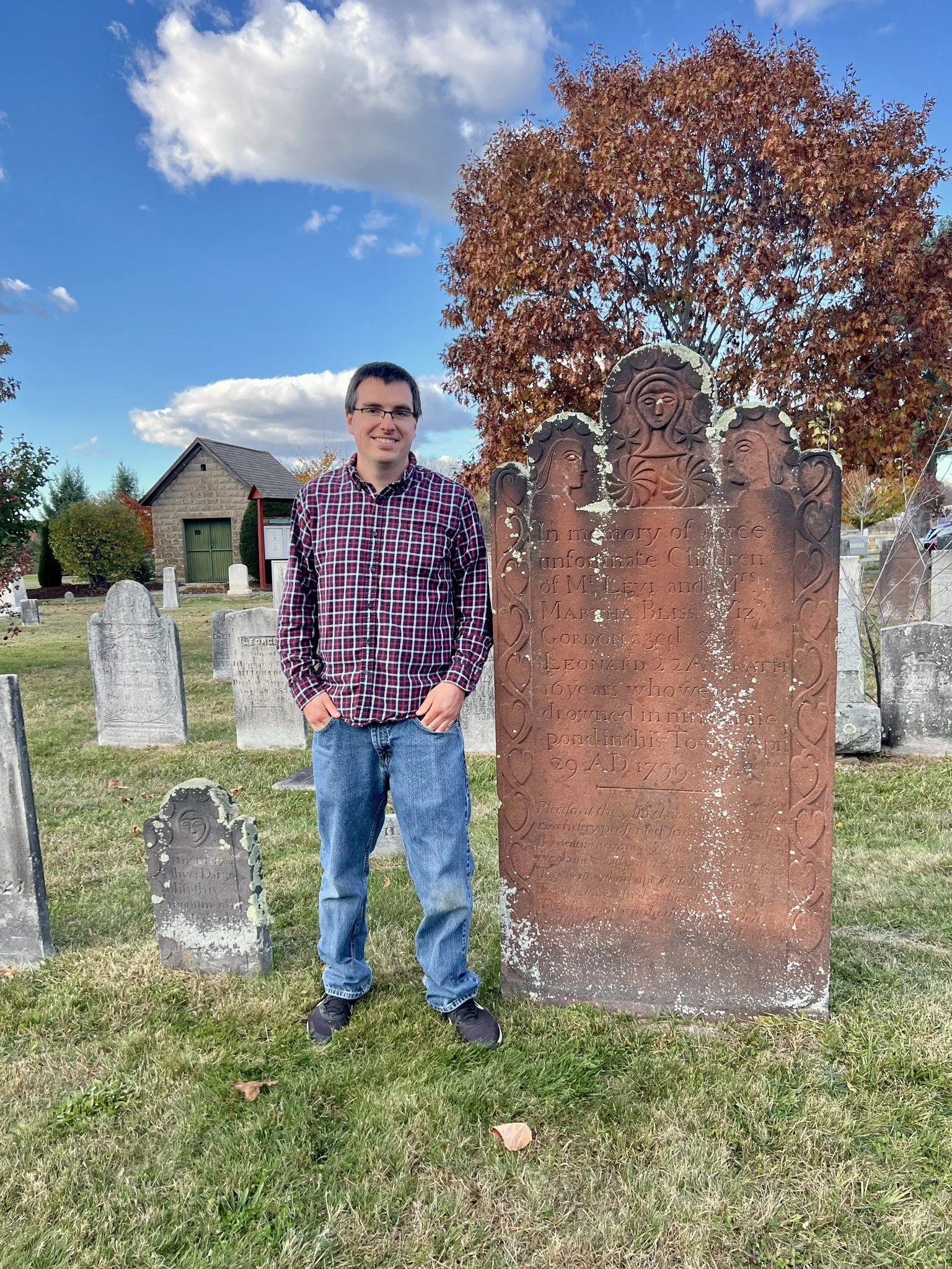 A young man standing in a cemetery next to a large, old gravestone with carved decorations and text. The gravestone appears weathered and has white lichen growth. Multiple other gravestones are visible in the background, with a tree with autumn-colored leaves behind him. The sky is partly cloudy with blue sky and white clouds.