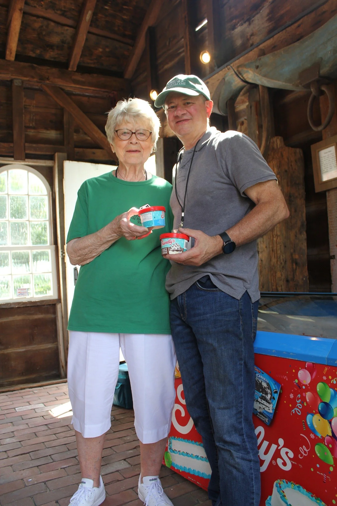 An elderly woman and a middle-aged man standing together inside a rustic wooden building, holding containers of ice cream. There is a colorful ice cream freezer in the background.