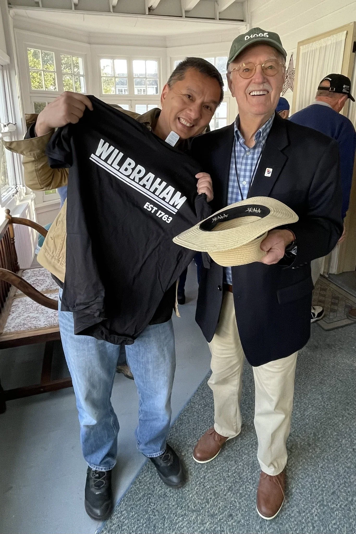 Two men standing indoors, one holding a Wilbraham jacket and a large straw hat, smiling at the camera. The man on the left wears a tan jacket and jeans, the man on the right wears a dark blazer, light-colored pants, and glasses.