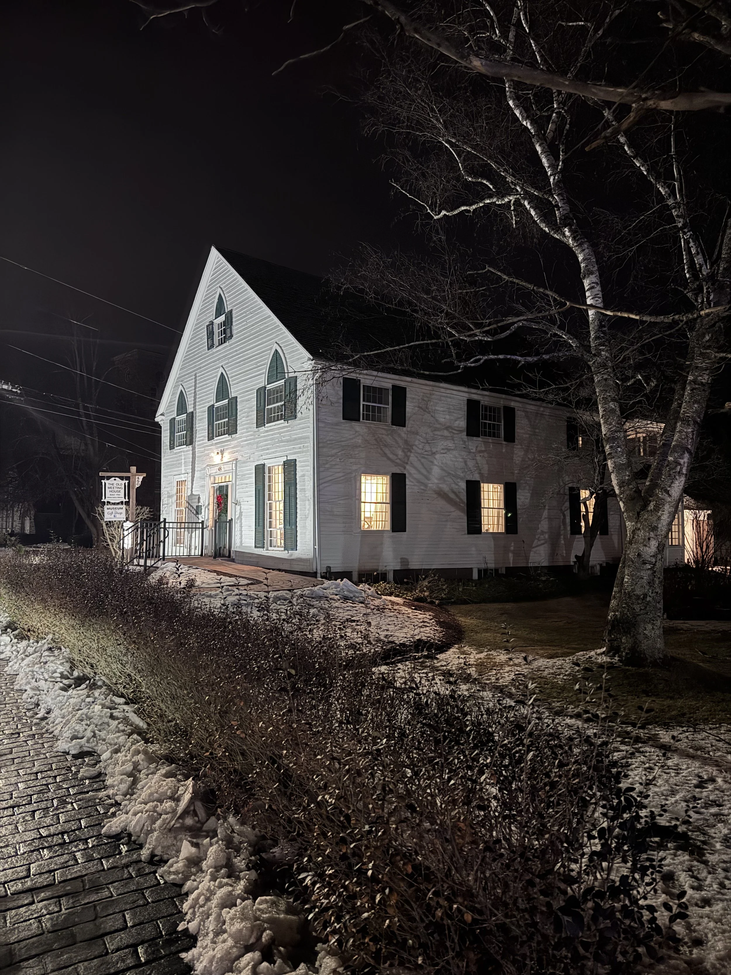 A white two-story house with black shutters and lights on inside, illuminated at night. The house has a steep roof and pointed arched windows on the upper floor. There is snow on the ground and a large bare tree to the right, with a brick pathway and some bushes in the foreground.