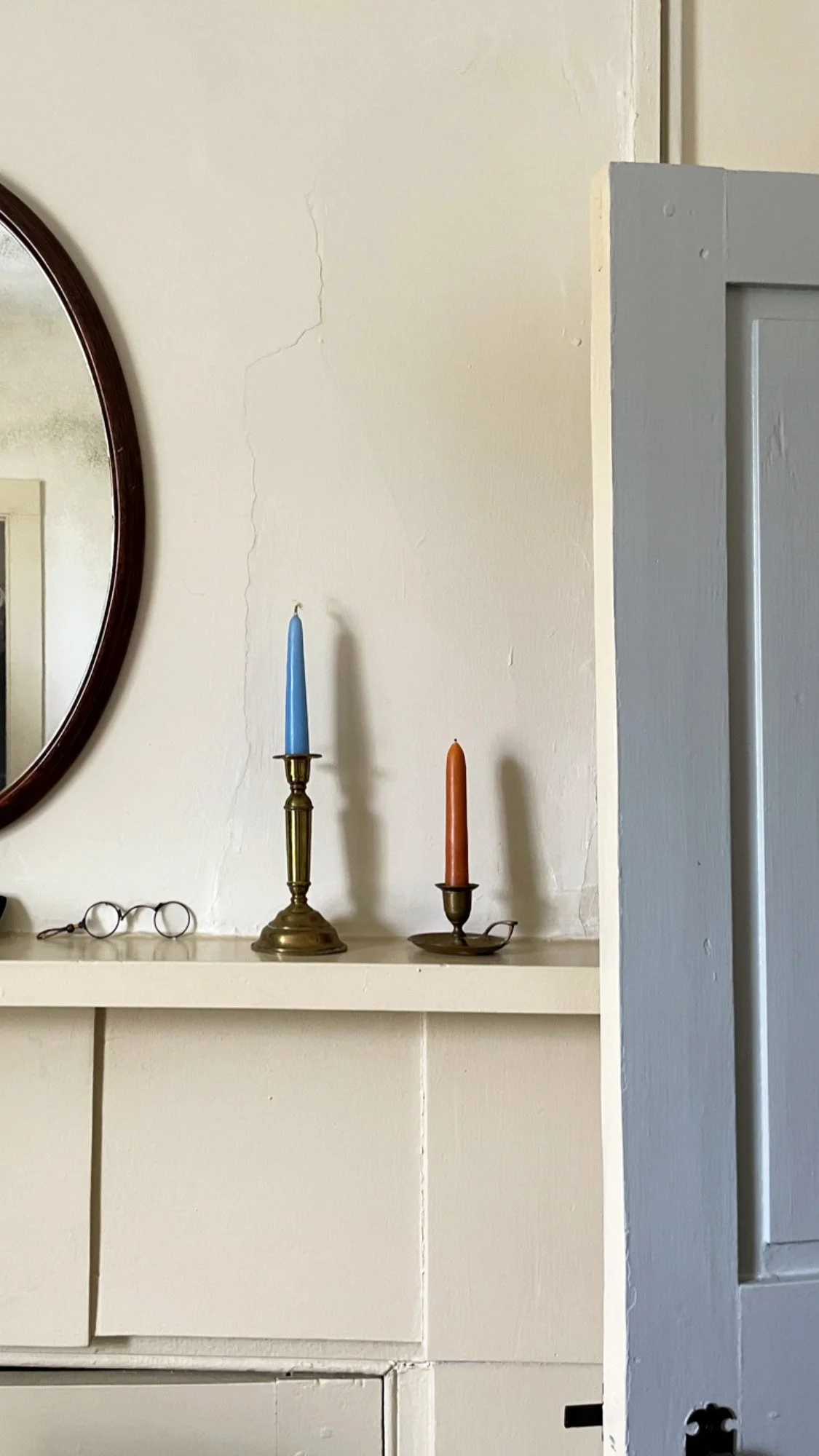 A beige wall with visible cracks, a portrait mirror, a white shelf with a pair of eyeglasses, and two candlesticks holding tall blue and orange candles next to a gray door frame.