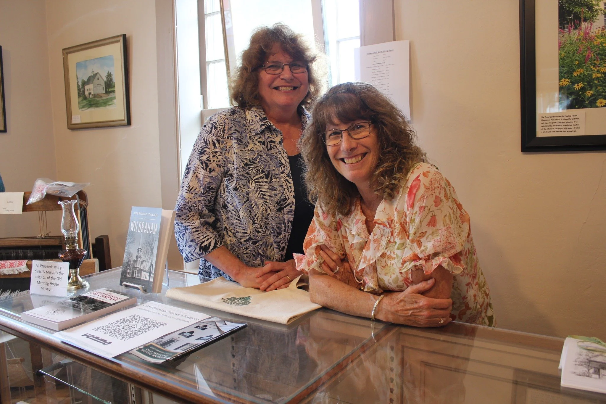 Two women smiling at a table in an art gallery, one standing and one leaning on the table, with framed artwork on the wall behind them.