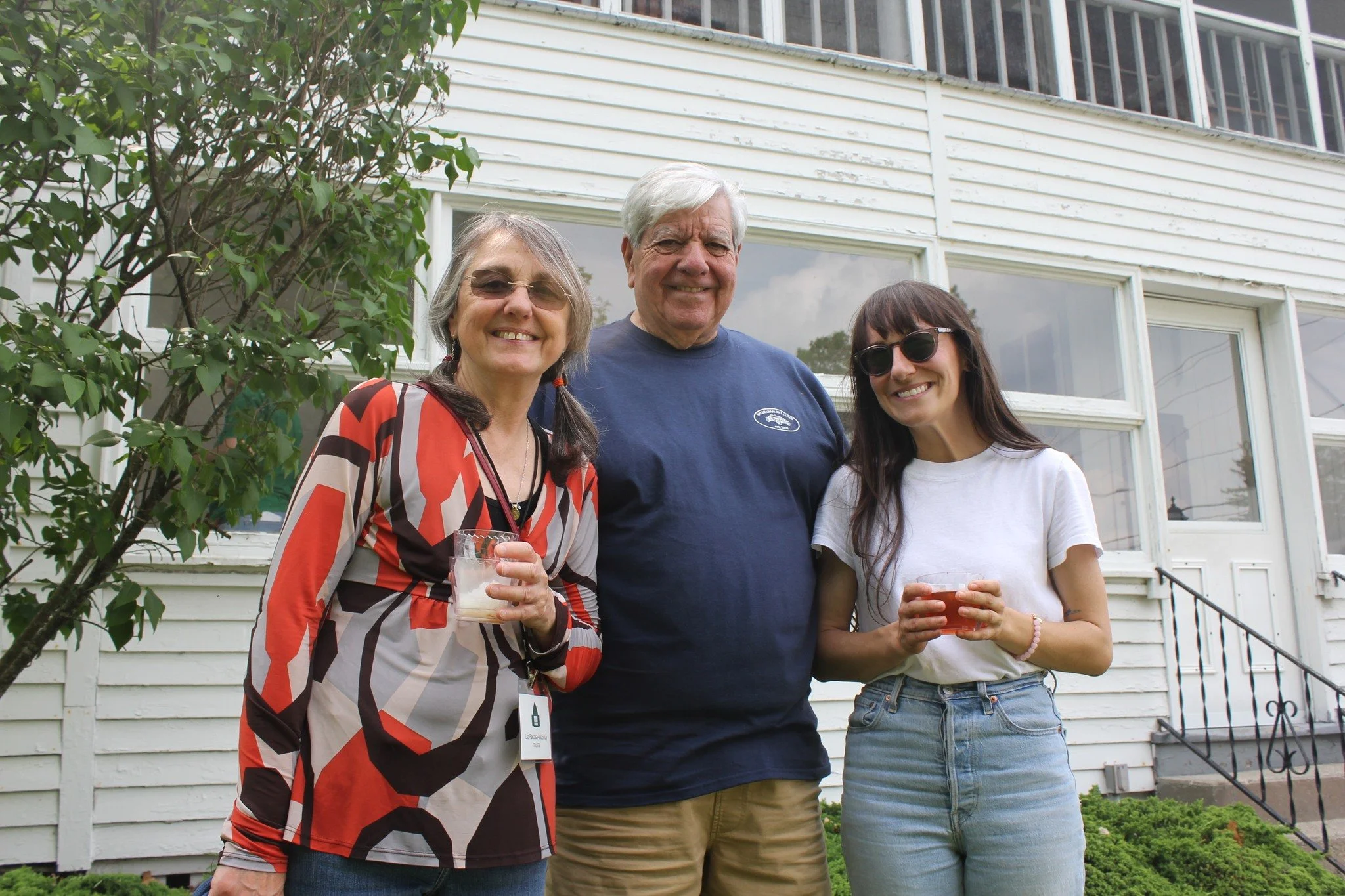Three people standing outside in front of a house, smiling, holding drinks, with a woman on the left wearing glasses and a patterned blouse, a man in the middle with white hair wearing a navy t-shirt, and a young woman on the right with dark hair, sunglasses, and a white t-shirt.