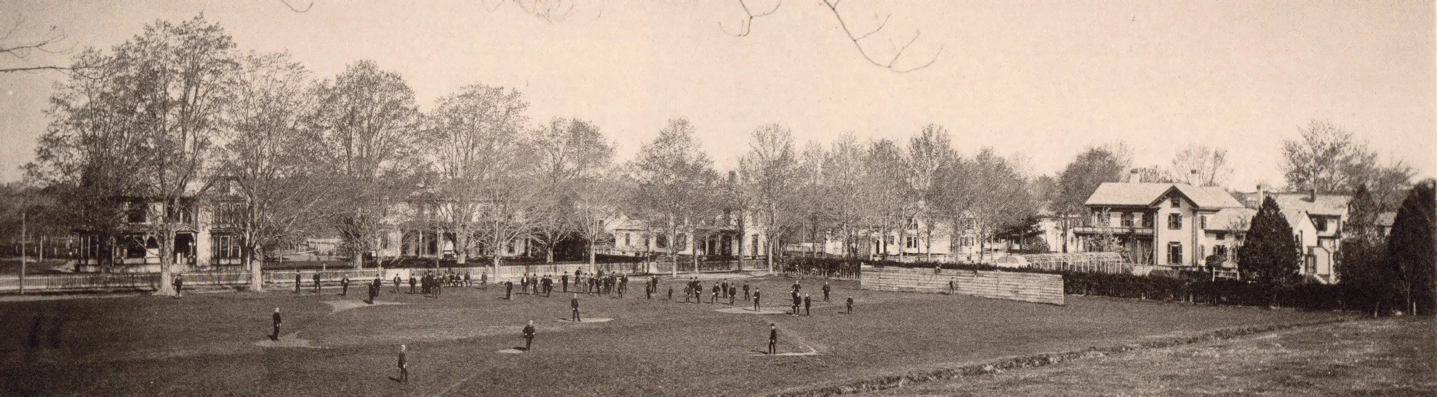 A vintage black-and-white photograph of a baseball game in a park with players on the field and houses in the background.