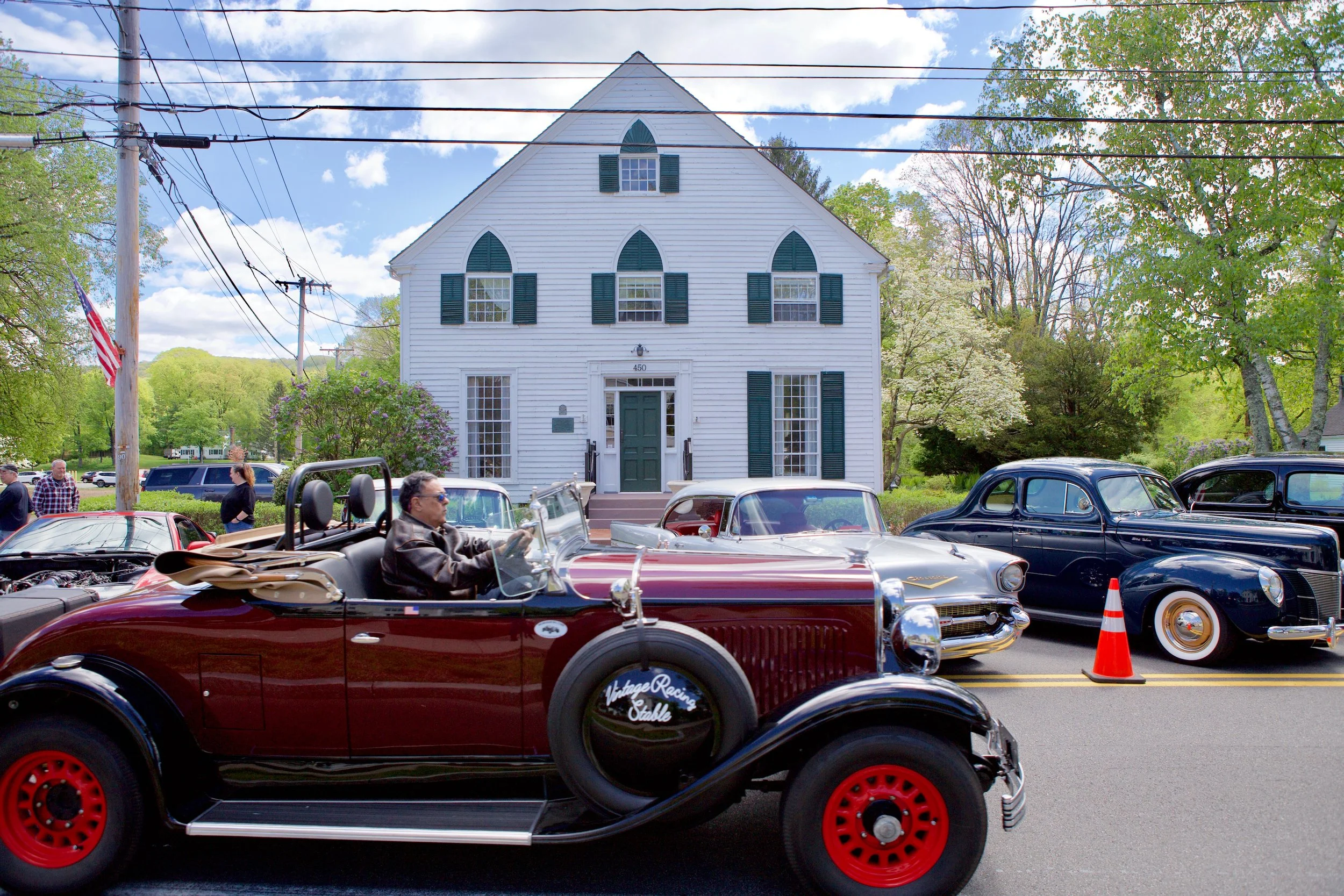 Vintage cars parked outside a white, historic church building with green shutters, under a partly cloudy sky and surrounded by green trees.