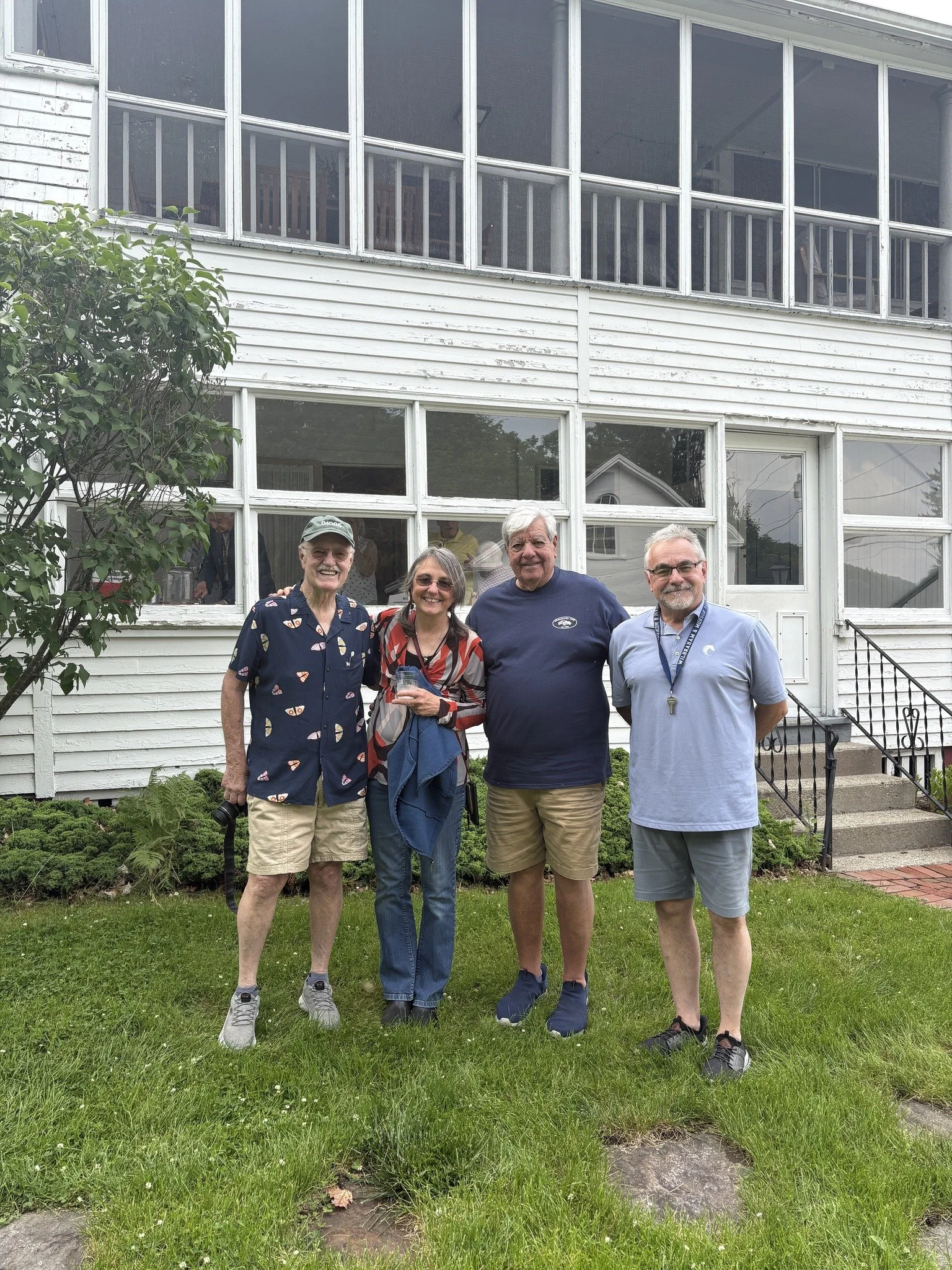 Four people standing outside in front of a white house, smiling, with a grassy yard and shrubbery.