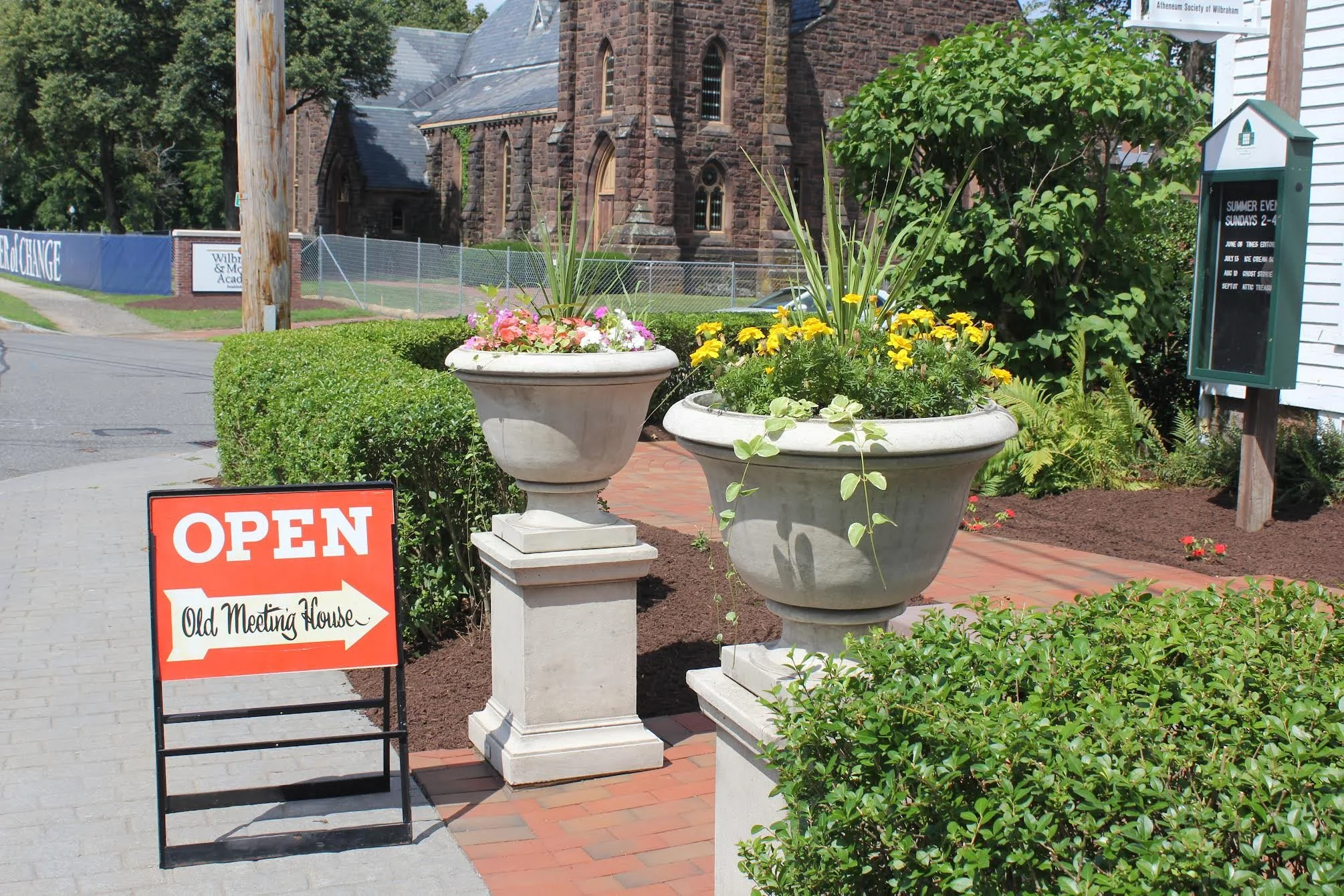 Sidewalk view with flower pots, bushes, and an orange 'Open' sign directing to Old Meeting House, outside a historic brick church, with a churchyard and informational sign in the background.