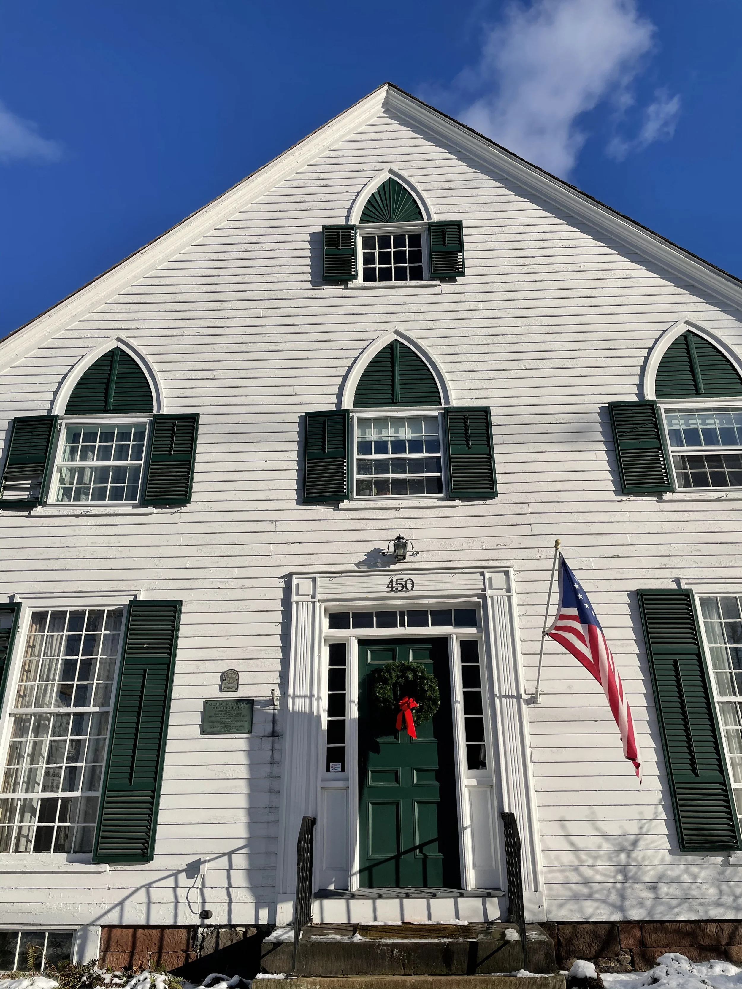 White wooden house with green shutters, resembling a church, decorated with a Christmas wreath and red bow on the green front door. An American flag is displayed outside. The house number is 450. The sky is clear and blue.