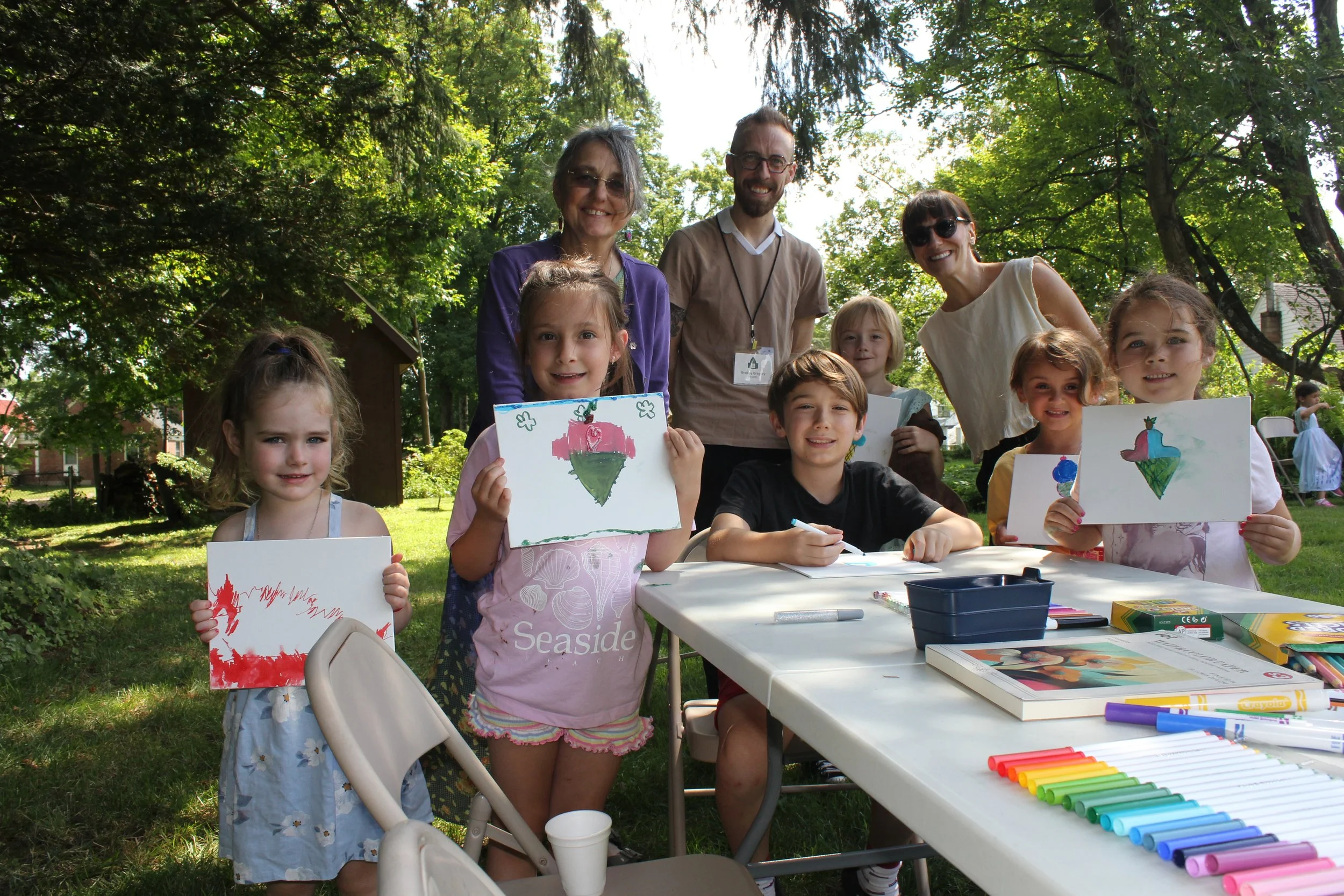 Children and adults outdoors at a craft activity table, showing their drawings of colorful ice cream cones with shades of pink, green, blue, and red, surrounded by green trees.