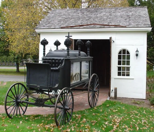 A black antique hearse with decorative finials on top, parked in front of a white small garage with a brick path leading inside, surrounded by grass and autumn leaves.