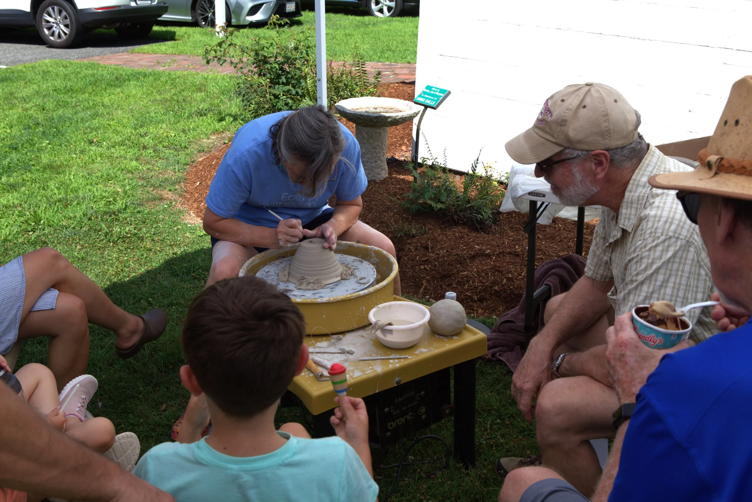 A woman is shaping a clay pot on a pottery wheel while people watch outdoors on a grassy area.