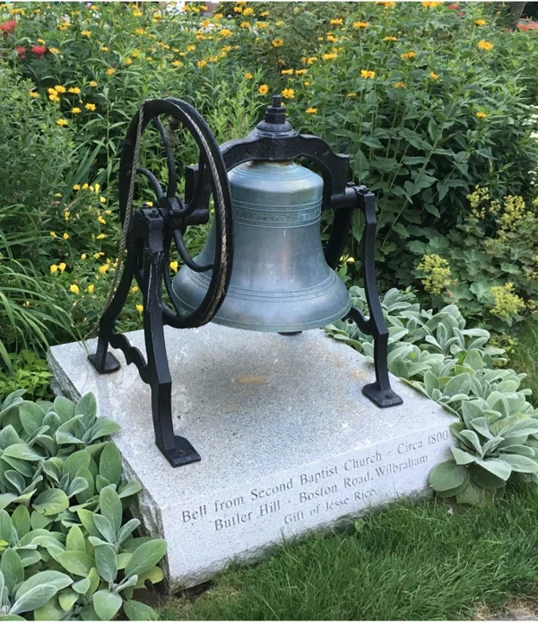 An antique bell mounted on a stone base surrounded by green plants and yellow flowers in a garden.